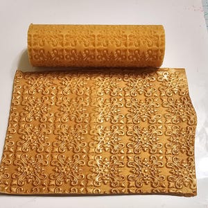 May include: A gold-colored rolling pin with a decorative pattern, used for creating embossed designs on fondant or dough. The rolling pin is shown next to a sheet of fondant with the same pattern embossed on it.