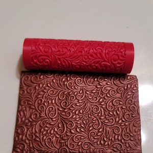 May include: A red rolling pin with a decorative floral pattern. The rolling pin is used to create an embossed design on fondant or clay. The image also shows a sample of the design created by the rolling pin.