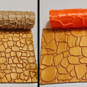 May include: Two silicone rolling pins with a stone pattern. The rolling pins are beige and orange. The beige rolling pin is on top of a gold textured surface. The orange rolling pin is on top of a gold textured surface.
