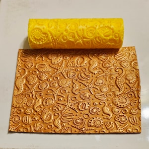May include: A yellow rolling pin with a seashell and starfish design. The rolling pin is used to create an embossed pattern on a gold-coloured surface.
