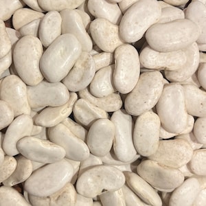 Puede incluir: Una imagen de primer plano de un montón de frijoles blancos secos. Los frijoles son lisos y de forma ovalada.