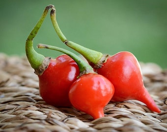 May include: Three red chili peppers with green stems, sitting on a woven brown surface.