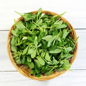 Pode incluir: Uma cesta de vime cheia de folhas frescas de feno-grego verde. A cesta está sobre uma superfície de madeira branca.