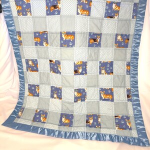 Puede incluir: Un edredón de retazos con un patrón repetido de corgis. El edredón presenta cuadrados en tonos de azul y gris, con un borde de satén azul. Los corgis se muestran en diferentes poses sobre un fondo azul.