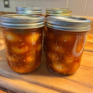 Puede incluir: Cuatro frascos de vidrio llenos de un líquido rojizo-anaranjado y orbes blancos, probablemente huevos en escabeche. Los frascos tienen tapas plateadas y están sobre una tabla de cortar de madera. El contenido es visible a través del vidrio transparente.