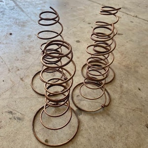 May include: Two antique metal springs with a spiral design. The springs are a rusty brown color and stand upright on a concrete surface. Each spring has a circular base and a series of coils that ascend upwards.