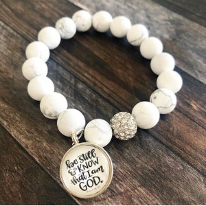 以下が含まれることがあります： 白いビーズのブレスレットに、"Be still & know that I am GOD"と書かれたシルバーのチャームが付いています。チャームは、ラインストーンが付いたシルバーのビーズに繋がっています。