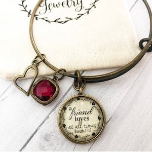 May include: A gold bangle bracelet with three charms: a heart, a red gemstone, and a round charm with the text "a friend loves at all times Proverbs 17:17".