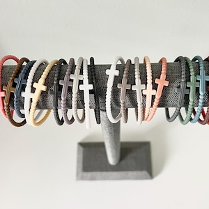 以下が含まれることがあります： 十字架のデザインが施されたカラフルなシリコンブレスレットのコレクション。ブレスレットは灰色の表面に一列に並んでいます。