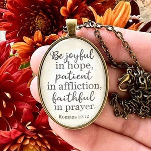 May include: A bronze necklace with an oval pendant featuring the Bible verse "Be joyful in hope, patient in affliction, faithful in prayer. Romans 12:12".