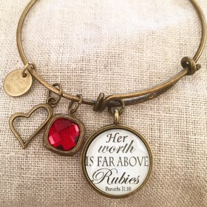 May include: A gold bangle bracelet with charms. The charms include a heart, a red gemstone, and a round charm with the text "Her worth is far above rubies Proverbs 31:10".