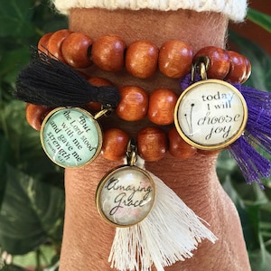 May include: A wooden beaded bracelet with three charms. The charms have inspirational messages: "But the Lord stood with me and gave me strength", "Today I will choose joy", and "Amazing Grace". Each charm has a tassel.
