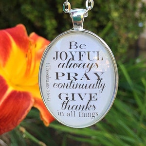 May include: A silver necklace with a white oval pendant that says "Be Joyful always Pray continually Give thanks in all things 1 Thessalonians 5:16-18".