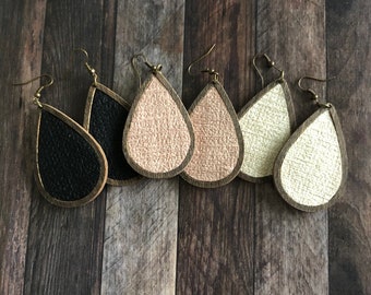 May include: A selection of teardrop earrings in black, rose gold, and cream. Each earring has a textured finish, a gold-coloured frame, and a hook. The earrings are arranged on a dark wood grain surface, showcasing their different colours.
