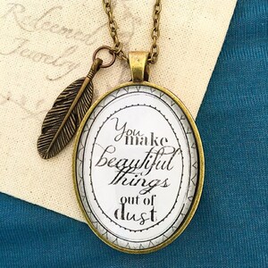 May include: A gold necklace with a round pendant featuring the text "You make beautiful things out of dust". The pendant is attached to a chain with a small feather charm.