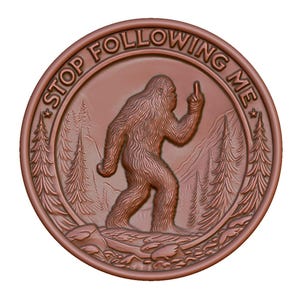 May include: A round, copper-colored coin with a raised image of a Bigfoot giving the middle finger. The coin has the words "Stop Following Me" around the edge.