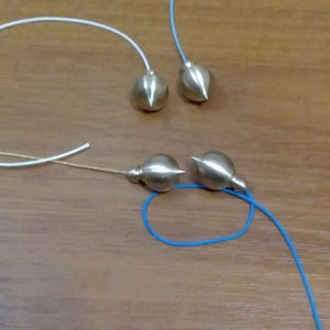 May include: Four brass pendulums with different colored cords. Each pendulum has a spherical body with a pointed tip. The cords are white, blue, and tan. The pendulums are arranged on a wooden surface.