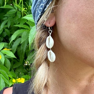 May include: A pair of dangle earrings featuring two white cowrie shells stacked vertically on each earring. The shells are connected by thin silver-toned wire, which also forms the hook mechanism for the earlobe attachment.