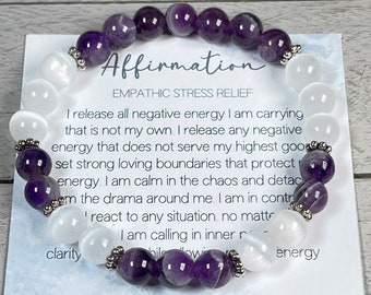 May include: Amethyst and white moonstone bracelet with silver beads. The bracelet is on a white card with the text "Affirmation Empathic Stress Relief" and a quote about releasing negative energy. The card also says "A Moonlit Canary".