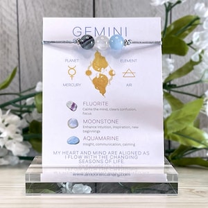 May include: A gray string bracelet with three beads: a gray fluorite, a white moonstone, and a light blue aquamarine. The bracelet is displayed on a white card with the text "GEMINI" and information about the stones and their astrological meanings.