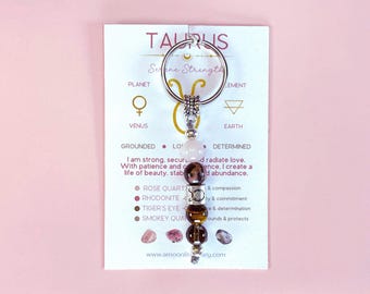 May include: A Taurus keychain with a silver keyring and a charm featuring various gemstone beads. The card behind the charm has the word "TAURUS" at the top, with text about the zodiac sign and its associated elements.
