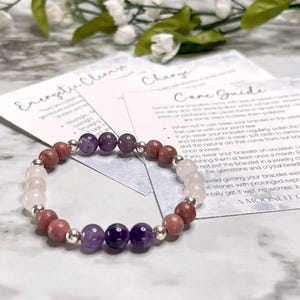May include: A beaded bracelet featuring amethyst, rose quartz, and rhodochrosite gemstones, interspersed with silver-colored beads. The bracelet is displayed with care instructions and a floral background.