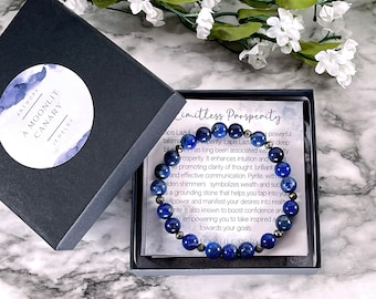 Puede incluir: Un brazalete azul y plateado con cuentas de lapislázuli y pirita. El brazalete está en una pequeña caja con una tarjeta blanca que dice "Prosperidad ilimitada" e incluye una descripción de las piedras y sus propiedades.