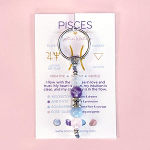 May include: A silver Pisces zodiac keychain with a crystal charm. The charm features amethyst, aquamarine, moonstone, and rose quartz crystals. The card reads "Pisces, Mystic Waters, Planet Jupiter Neptune, Element Water, Creative Dreamy Gentle, I flow with the...se in love and trust. My heart is...n. my intuition is clear, and my cr...ly is in the flow. Moonstone...tion & dreams, Amethyst...& protective, Aquamarine...tional flow & courage, Rose Quar...ving and gentle, www.amooanary.com".