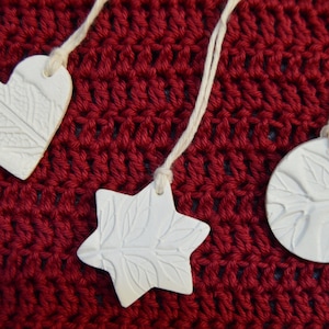 May include: Three white ceramic ornaments with leaf patterns hang against a red crocheted background. The ornaments are a heart, a star, and a circle, each with a string for hanging. The ornaments are a neutral colour.