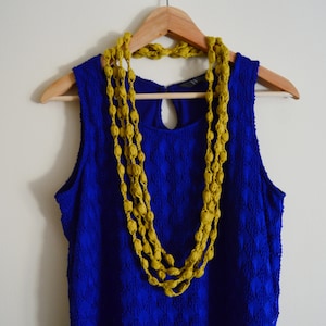K&ouml;nnte beinhalten: Ein blaues, &auml;rmelloses Kleid mit einer gelben Perlenkette, die an einem Holzb&uuml;gel h&auml;ngt.