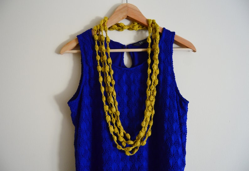 K&ouml;nnte beinhalten: Ein blaues, &auml;rmelloses Kleid mit einer gelben Perlenkette, die an einem Holzb&uuml;gel h&auml;ngt.