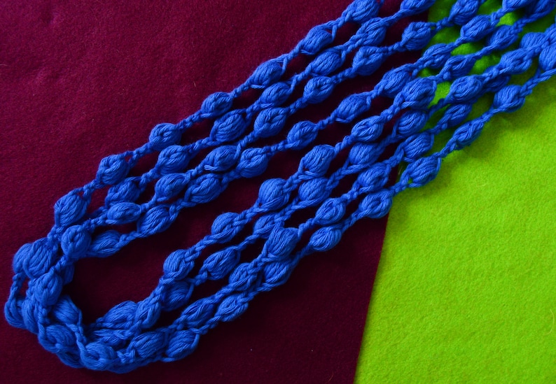 K&ouml;nnte beinhalten: Nahaufnahme einer blauen geh&auml;kelten Halskette aus mehreren Garnstr&auml;ngen. Die Halskette besteht aus kleinen, runden Perlen, die in einem sich wiederholenden Muster miteinander verbunden sind. Die Halskette liegt auf einer roten und gr&uuml;nen Oberfl&auml;che.