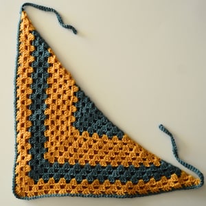 Puede incluir: Un chal triangular de ganchillo con un patrón de color amarillo mostaza y azul verdoso.