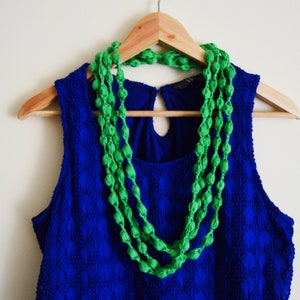 K&ouml;nnte beinhalten: Ein blaues, &auml;rmelloses Kleid mit einer gr&uuml;nen Perlenkette, die an einem Holzb&uuml;gel h&auml;ngt.