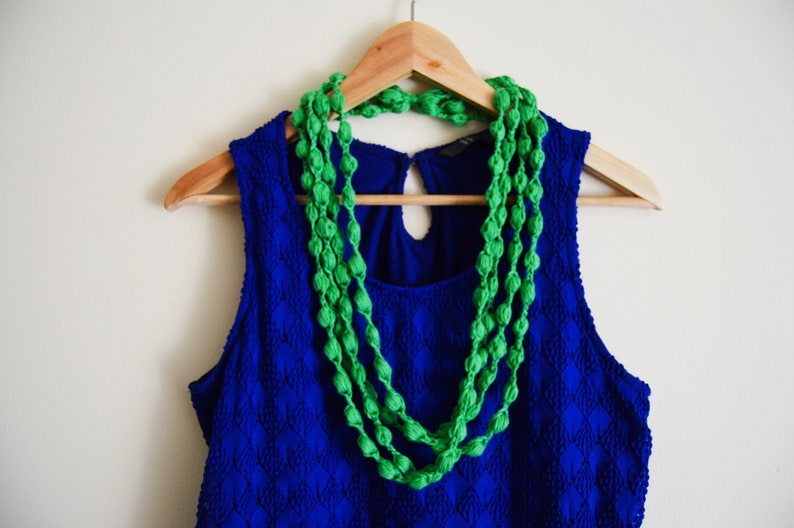 K&ouml;nnte beinhalten: Ein blaues, &auml;rmelloses Kleid mit einer gr&uuml;nen Perlenkette, die an einem Holzb&uuml;gel h&auml;ngt.