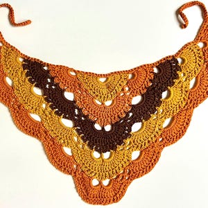 May include: A handmade crochet shawl in shades of orange, yellow, and brown. The shawl has a scalloped edge and a fan-like pattern. It features two tie strings at the top for securing around the neck.