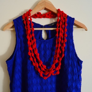 K&ouml;nnte beinhalten: Ein blaues &auml;rmelloses Kleid mit einer roten geh&auml;kelten Halskette. Das Kleid hat ein Rautenmuster und die Halskette besteht aus mehreren Reihen roter Bommeln.