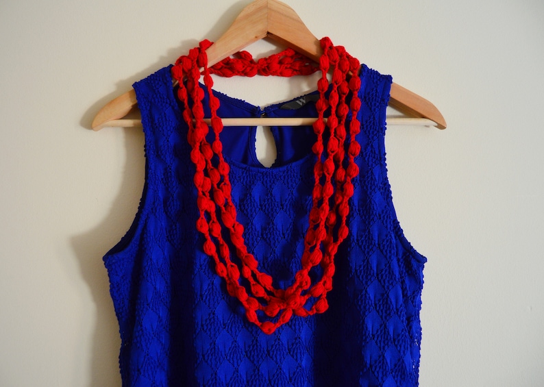 K&ouml;nnte beinhalten: Ein blaues &auml;rmelloses Kleid mit einer roten geh&auml;kelten Halskette. Das Kleid hat ein Rautenmuster und die Halskette besteht aus mehreren Reihen roter Bommeln.