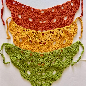 May include: Three handmade crochet neck scarves in shades of orange, yellow, and green. Each scarf features a scalloped edge and a series of circular cutouts. The scarves have tie closures at the ends.