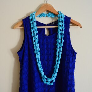 K&ouml;nnte beinhalten: Ein blaues, &auml;rmelloses Kleid mit einer blauen Perlenkette, die an einem Holzb&uuml;gel h&auml;ngt.
