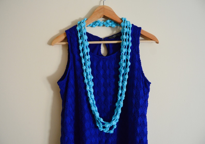 K&ouml;nnte beinhalten: Ein blaues, &auml;rmelloses Kleid mit einer blauen Perlenkette, die an einem Holzb&uuml;gel h&auml;ngt.