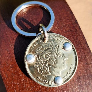 Könnte beinhalten: Ein goldfarbener Münzschlüsselanhänger mit einem silbernen Schlüsselring. Die Münze zeigt das Profil einer Person mit lockigem Haar und griechischem Text. Drei silberne Nieten sind am Rand der Münze angebracht. Der Schlüsselanhänger liegt auf einer Holzoberfläche.