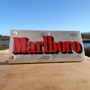 Può includere: Un cartello metallico rettangolare con la scritta "Marlboro" in rosso, con lettere in stile 3D. Il cartello è argentato con quattro bulloni argentati in ogni angolo. Lo sfondo è una superficie di legno con cielo blu.