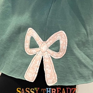 May include: A teal green sweatshirt with a pink bow applique on the back. The bow has a white floral pattern and is outlined with white stitching. The text "SASSY THREADZ" and "4"-"10" TALL" is printed on the bottom of the sweatshirt.
