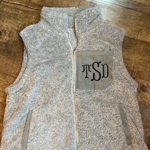 May include: A gray sherpa vest with a zipper closure and a brown patch pocket on the front. The pocket has the monogram "TSD" embroidered on it.
