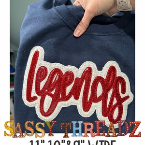Könnte beinhalten: Ein marineblauer Sweatshirt mit einem roten und weißen Pailletten-Applikationen "Legends". Der Sweatshirt ist in den Größen 11", 10" und 9" breit erhältlich. Der Text "SASSY THREADZ" ist unterhalb der Applikation gedruckt.