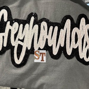 Op de afbeelding: Het woord "Greyhound" gespeld in witte letters met een zwarte omtrek en een glinsterende afwerking op een grijs T-shirt.