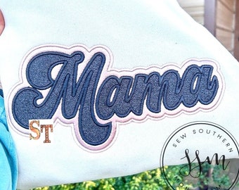 May include: Cream-colored sweatshirt with the word "Mama" embroidered in a navy blue, glittery script with a light pink outline. A small "ST" patch is visible below the word. The sweatshirt is from "Sew Southern Moms".