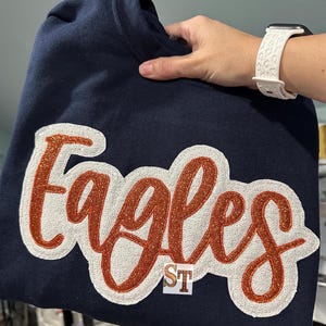 Puede incluir: Sudadera azul marino con una aplicación de "Eagles" de purpurina blanca y naranja.