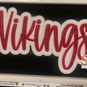 May include: A red and white embroidered patch with the word "Vikings" in a cursive font. The patch is on a black fabric background.